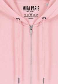 Hoodie zippé en coton rose avec une capuche à cordon, une fermeture éclair argentée et une étiquette de marque avec des instructions d'entretien à l'intérieur.