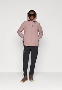Lichtroze hoodie-jack met zwarte accenten, ritszakken en verstelbare trekkoorden, gecombineerd met zwarte cargobroek en bruine wandelschoenen.