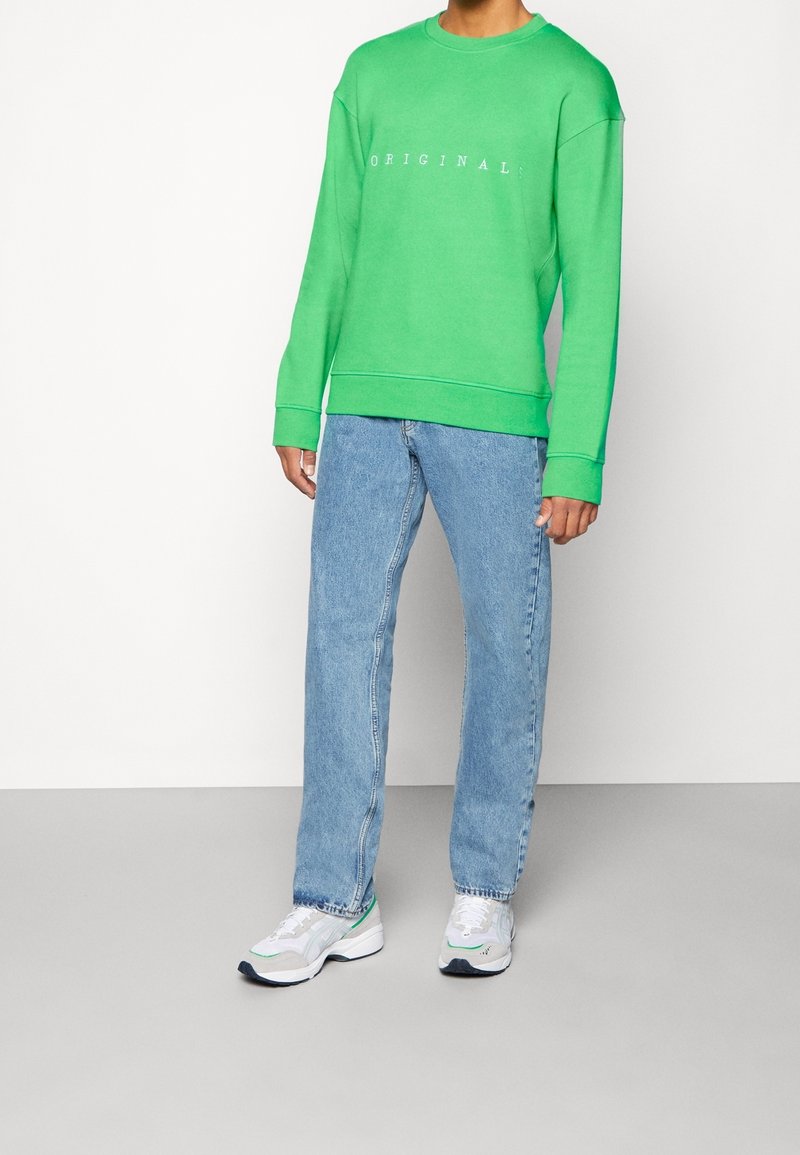 Grön sweatshirt med texten "ORIGINAL" i vitt, i kombination med ljusblå denimjeans. Vita sneakers med grå och gröna detaljer.