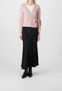 Una donna che indossa un cardigan avvolgente rosa pallido sopra a una maglietta bianca, abbinato a una lunga gonna nera e stivaletti neri, in piedi su uno sfondo semplice.