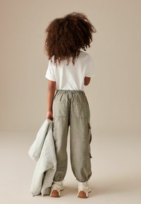 Kind trägt ein weißes T-Shirt und graue Cargo-Hosen mit elastischen Bündchen, hält einen hellgrauen Sweatshirt und Schuhe mit weißen und beige Akzenten.