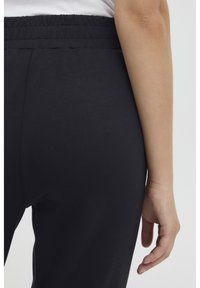Svarte stretchy bukser med elastisk midjebånd, glatt tekstur og en tettsittende design. Modellens hånd hviler på hoften.