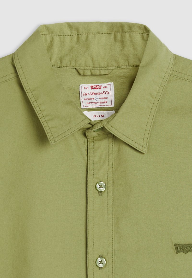 Chemise verte claire ajustée, réalisée en tissu doux, avec un col pointu, deux boutons et un petit logo de marque dans le coin.