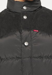 Giubbotto puffer nero con accento in corduroy sul colletto, bottoni a pressione argentati e etichetta logo rossa. Superficie testurizzata con design trapuntato.