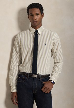 Koszula w paski zapinana na guziki w bladym żółtym i białym kolorze, zestawiona z ciemnym dzianinowym krawatem. Ciemne jeansy i brązowy pasek ze srebrną klamrą.