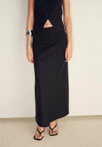 Schwarze Maxikleid aus glattem, fließendem Stoff. Kombiniert mit einem enganliegenden schwarzen Cropped-Top. Einfache schwarze Sandalen ergänzen das Outfit.