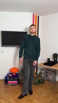 Σκούρο πράσινο πουκάμισο κοκκόρι με κουμπιά και τσέπη στο στήθος, γκρι τζιν με ίσια πόδια και μαύρα γυαλιστερά παπούτσια· εσωτερικός χώρος με πολύχρωμη διακόσμηση.