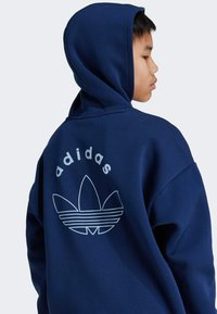 Sudadera con capucha de color azul marino que presenta un gran logo blanco de Adidas en la parte posterior, con tres franjas y texto redondeado arriba del logo.