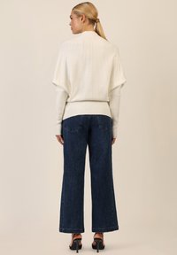 Weißer gerippter Strickpullover mit hohem Kragen, kombiniert mit dunkelblauen weit geschnittenen Jeans mit dezenten strukturierten Details und schwarzen Absatzsandalen.