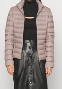 Doudoune matelassée mauve clair avec un col haut et une fermeture éclair, portée sur un col roulé noir et une jupe en cuir noir avec des boutons.