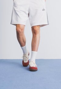 Pantaloni scurți atletici albi cu dungi negre pe laterale, asortați cu teniși roșii și albi și șosete albe până la glezne, prezentate pe un fond albastru.