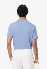Polo shirt azzurro chiaro a maniche corte con colletto. Tessuto morbido con vestibilità rilassata. Indossato con pantaloni bianchi e reggendo una giacca di colore chiaro.