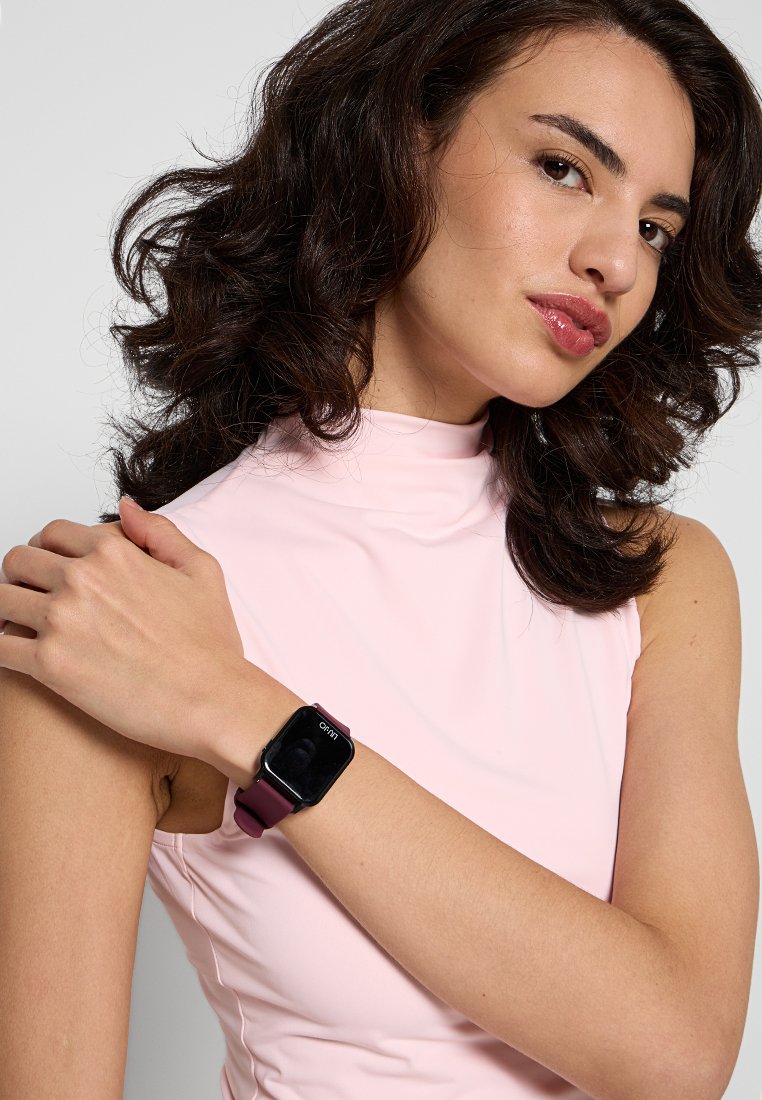 Smartwatch mit einem quadratischen schwarzen Zifferblatt und einem bordeauxfarbenen Silikonarmband. Modell trägt ein pinkes ärmelloses Oberteil und zeigt die Uhr an ihrem Handgelenk.