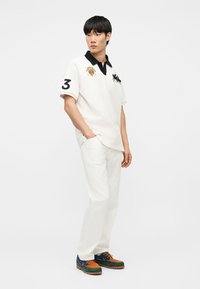 Άνδρας που φοράει λευκό πόλο μπλουζάκι με μαύρο γιακά και αριθμό 3 στο μανίκι, λευκό παντελόνι και πολύχρωμα παπούτσια με κορδόνια, στέκεται με τα χέρια στις τσέπες.