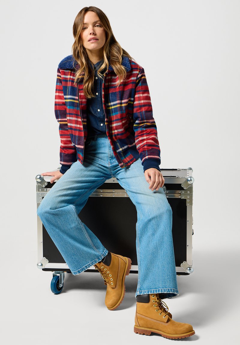 Koszula w kratę w kolorach czerwonym, niebieskim i żółtym, z niebieskim kołnierzykiem, zestawiona z ciemnoniebieską koszulą, jasnoniebieskimi spodniami z szerokimi nogawkami i beżowymi botkami.