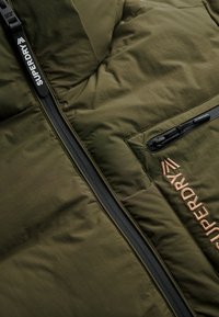 Gewatteerd jack in olijfgroen, met gewatteerde textuur, ritssluiting aan de voorkant met branding en een contrasterende zwarte ritssluitingzak.