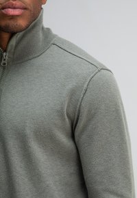 Grijze zip-up trui met een geribde kraag en een gladde textuur, met een op maat gemaakte pasvorm en strakke lijnen, die een modern ontwerp laat zien.