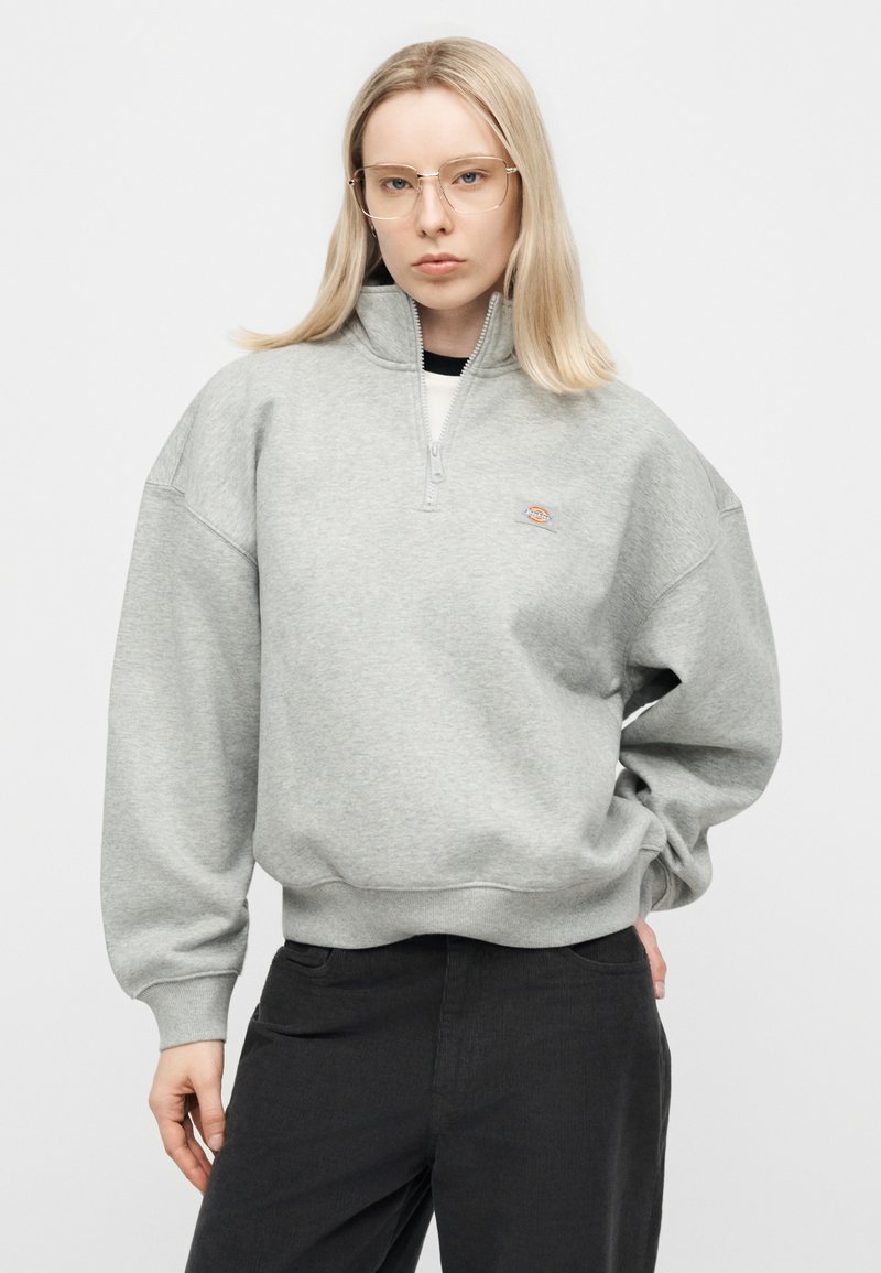 Donna con capelli biondi lisci, occhiali trasparenti, felpa grigia chiara con zip e pantaloni neri, in piedi con una mano in tasca.