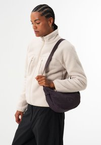 Leichte, dunkel lila Schultertasche mit einer runden Form und einer weichen Textur, ausgestattet mit einem einfachen Logo und einem verstellbaren Gurt.