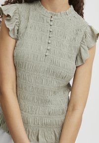 Haut vert clair, texturé, avec une manche courte à volants, un col rond et quatre boutons en tissu recouverts sur le devant. Design froissé.