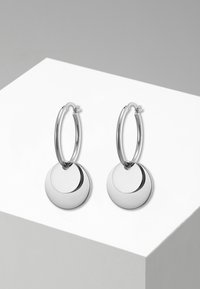 Gli orecchini a cerchio in argento presentano un pendente circolare lucido, una texture liscia e una finitura brillante, esposti su un piedistallo bianco.