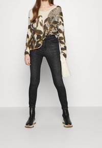 Strickpullover in Creme mit braunen und senfgelben Blumenmustern, kombiniert mit dunklen, hochgeschnittenen Jeans und schwarzen Stiefeletten. Hält eine weiße Handtasche.