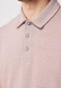 Lyserød polo skjorte lavet af blødt stof med en foldet krave, tre grå knapper og en brystlomme.