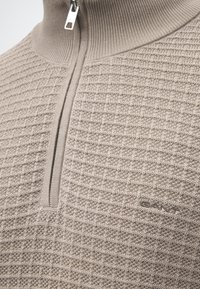 Pull beige en maille avec fermeture éclair quart, motif carré texturé et logo GANT brodé sur la poitrine.