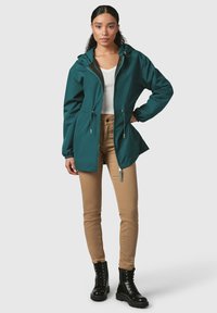 Giacca leggera turchese con cappuccio, cordini regolabili in vita e chiusura con zip. Abbinata a pantaloni beige e stivali neri con lacci.