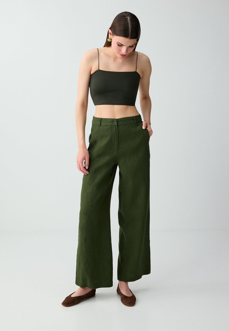 Femme portant un caraco court vert foncé et un pantalon large taille haute vert olive, debout devant un fond clair uni.