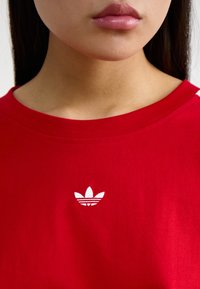 Sarkanais kokvilnas T-krekls ar apaļu kaklu. Uz krūtīm ir balts izšūts logotips ar trim lapām virs trim svītrām.