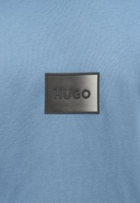Blauer Strickstoff mit einem schwarzen, rechteckigen Fleck in der Mitte, auf dem das Wort "HUGO" in Großbuchstaben geprägt ist.