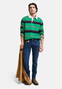 Camisa de rugby a rayas verdes con un cuello blanco, jeans azules y botas de ante marrón; sosteniendo una chaqueta marrón, posando frente a un fondo blanco liso.