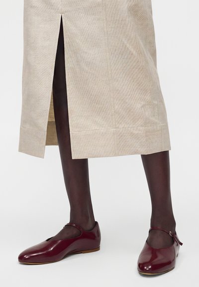 Πόδια με σκούρα καλσόν που φορούν μπορντό παπούτσια Mary Jane συνδυασμένα με μπεζ midi φούστα με σκίσιμο μπροστά.
