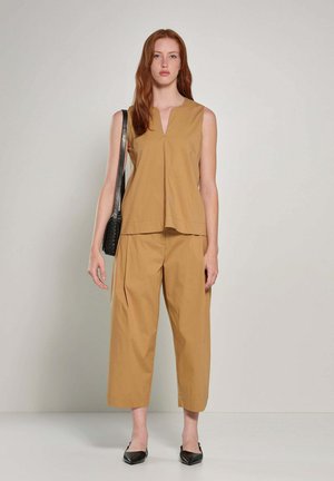 Kobieta z rudymi włosami, ubrana w beżowy, bezrękawnik i szerokie spodnie o długości do połowy łydki, czarne buty na obcasie z paskiem z tyłu oraz czarną torebkę na ramię.