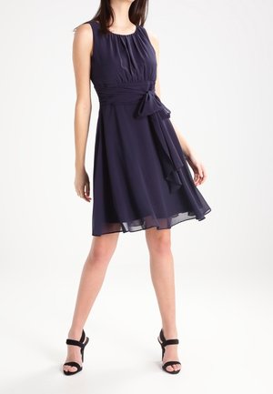 Vrouw draagt een mouwloze marineblauwe jurk tot over de knie met een tailleceintuur, gecombineerd met zwarte sandalen met hoge hakken, staand met gespreide benen.