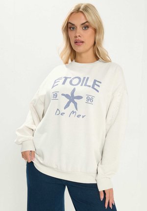 Lyshåret kvinde iført en løs hvid sweatshirt med en blå søstjerne og teksten "ÉTOILE 1996 De Mer" kombineret med mørkeblå jeans.