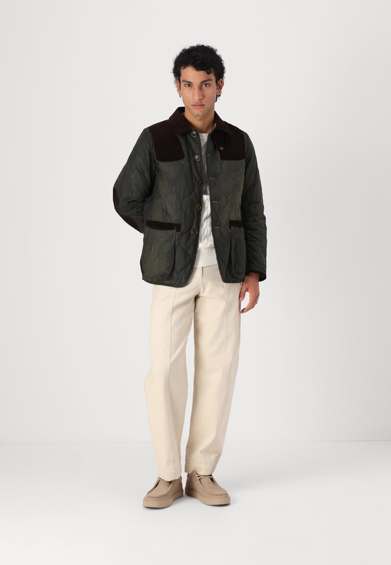 Gestepptes olivgrünes Jäckchen mit braunem Kragen und Taschen, Knopfleiste, getragen über einem hellen Pullover, kombiniert mit beigen Hosen und Schuhen.