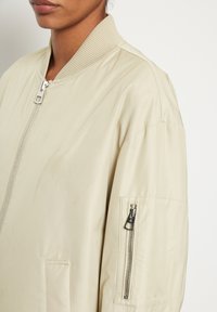 Beige bomberjack met geribbelde kraag, ritssluiting aan de voorkant en zijzak. Heeft een ritszak op de bovenarm voor extra functionaliteit.