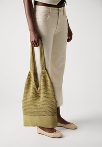 Sac fourre-tout en crochet vert olive avec de longues anses, design texturé et fond solide. Associé à un jean crème et des ballerines beige clair.