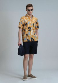 Bloemige kortekoepshirt in mosterdkleur met blauwe en zwarte accenten, gecombineerd met marineblauwe shorts en grijze instapschoenen, met een marineblauwe hoed in de hand.