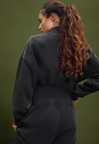 Felpa nera accorciata con maniche lunghe, tessuto testurizzato e vita elastica abbinata a pantaloni jogger coordinati. Sfondo verde uniforme.