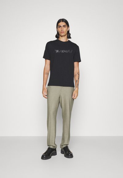 Black cotton t-shirt with a textured graphic design, paired with light grey trousers and black shoes featuring a thick sole and laces.