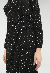 Robe noire cache-coeur avec des motifs de pois argentés métalliques, manches longues et un détail à nouer à la taille, fabriquée dans un tissu doux et texturé.