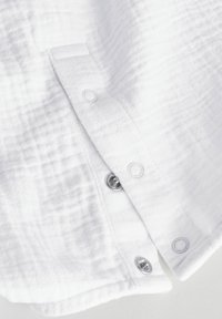 Camisa de algodón blanca con una superficie texturizada, que presenta botones a presión plateados y un dobladillo curvado en la parte inferior.