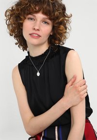 Schwarzes ärmelloses Oberteil mit einem Rüschen-Ausschnitt, das einen subtilen Glanz aufweist. Das Model trägt eine silberne Anhänger-Halskette über der Bluse.