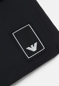 Tessuto nero con superficie strutturata, che presenta una toppa rettangolare con logo in bianco e nero, raffigurante un emblema stilizzato di aquila.