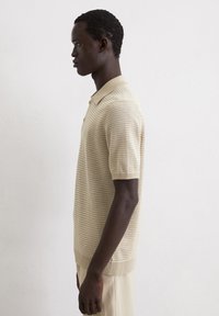 Beige knitted polo shirt with a textured pattern. Short sleeves and a ribbed collar. Side view shows relaxed fit and smooth fabric.