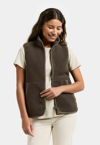 Glimlachende vrouw die een bruin fleecevest draagt over een beige shirt met korte mouwen en crèmekleurige broek, staand tegen een effen achtergrond.