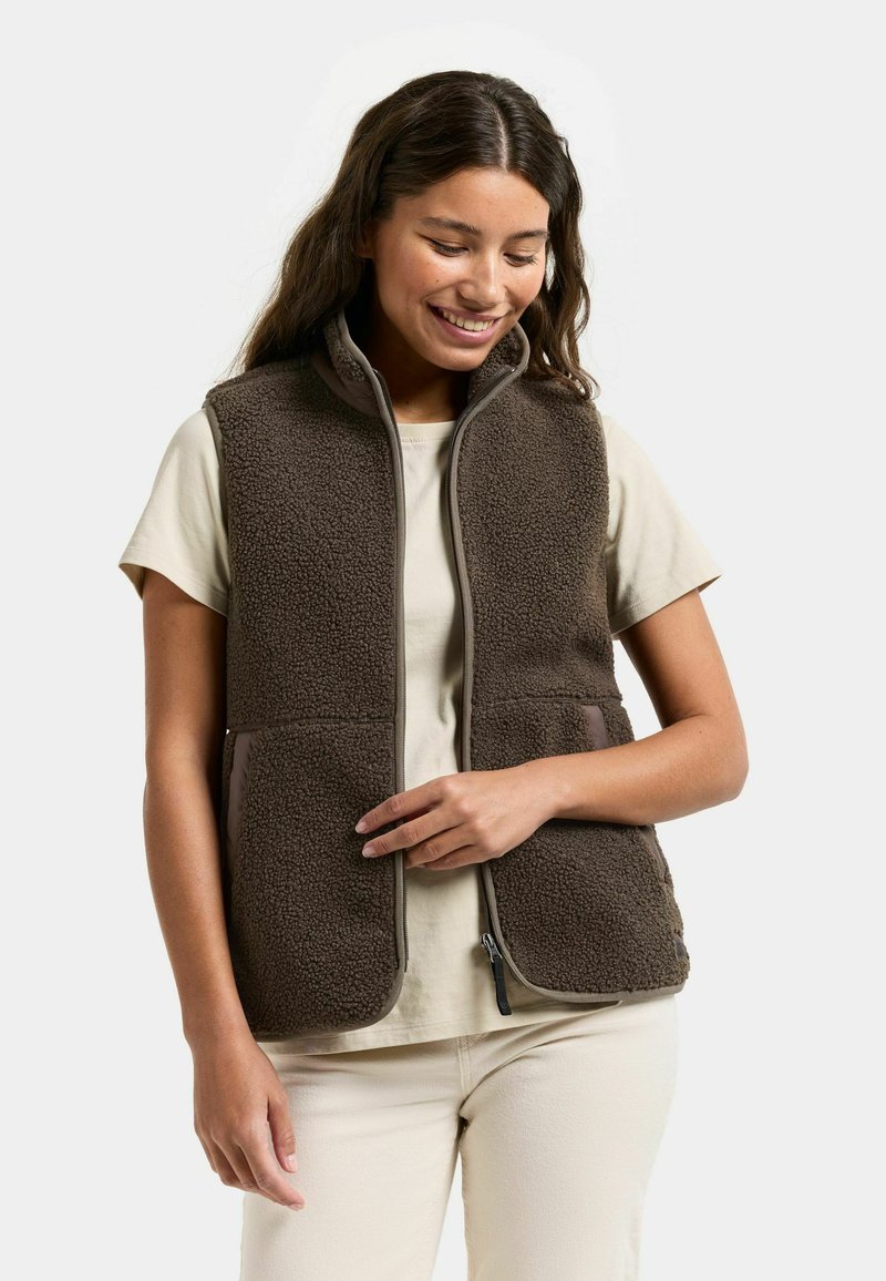 Glimlachende vrouw die een bruin fleecevest draagt over een beige shirt met korte mouwen en crèmekleurige broek, staand tegen een effen achtergrond.